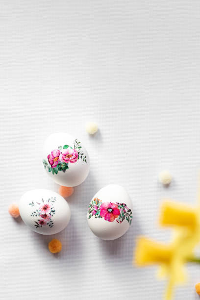 Three white eggs with floral patterns in shades of pink, red, and green on a white surface. Colorful pom-poms in orange, yellow, and white surround them. A blurred yellow flower is visible on the left. Perfect for spring or Easter decorations.