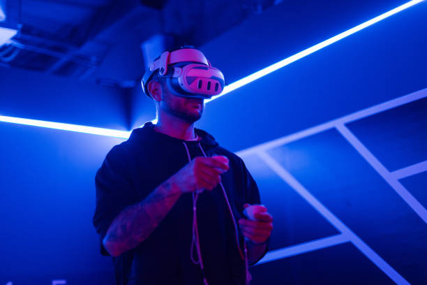istock Cool trendy man in vr glasses, watching 360 degree video with virtual reality headset on dark background with neon light. guy holds a joystick and plays virtual games 2201683507