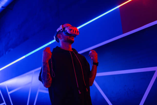 istock Cool trendy man in vr glasses, watching 360 degree video with virtual reality headset on dark background with neon light. guy holds a joystick and plays virtual games 2201687991