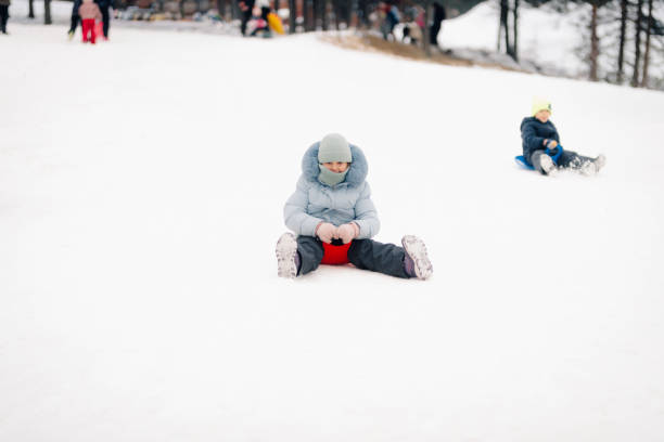 Happy little boy dressed in a green winter jacket, blue pants, black boots, and a red‑and‑black knit hat speeds downhill on a bright yellow sled in a snow‑covered forest. The background trees and snow are captured with intentional motion blur, emphasizing the sense of speed and excitement. Snow trails behind the sled add to the dynamic energy of the scene. The image conveys the thrill of sledding, childhood joy, and the beauty of outdoor winter recreation.Shot with Nikon D800