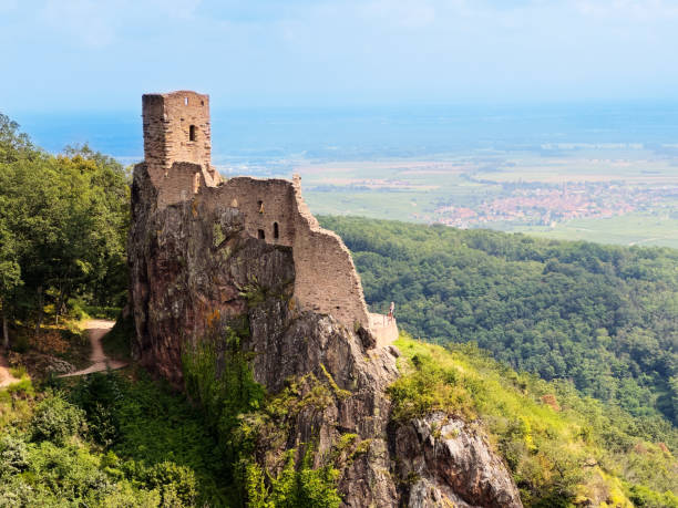 A medieval fortress ruin stands on a rocky hill, surrounded by lush green countryside and overlooking a village in the distance.