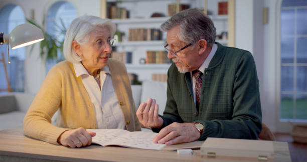 A senior Caucasian couple, dressed in cozy sweaters, sits at a wooden table in their home. They’re happily solving a crossword puzzle, fostering connection and mental engagement.