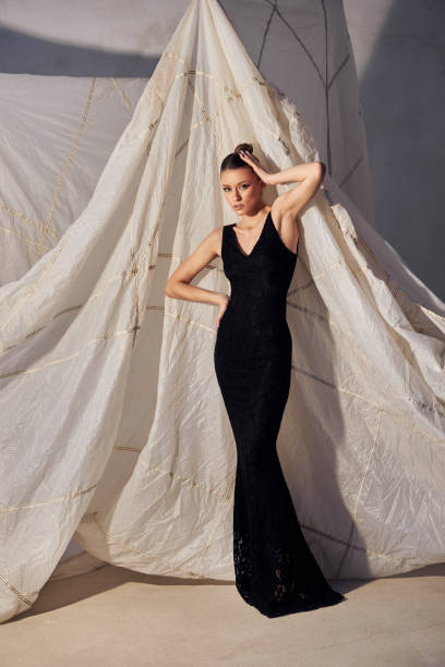 istock woman showcases a form-fitting black gown while elegantly posing with one hand on her hip. minimalistic background enhances the sophistication of the outfit, creating a striking silhouette 2201700654