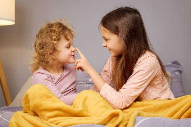 istock Two young girls share a joyful moment while sitting on a bed, playfully interacting with soft smiles and a cozy blanket around them, creating a warm and loving atmosphere at home 2201704697