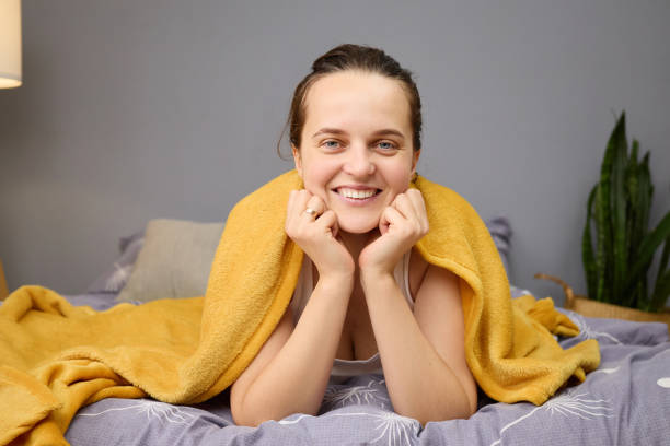 istock A woman is enjoying a peaceful moment at home, smiling while resting on a bed covered with a soft yellow blanket. She appears content and relaxed, showcasing a cozy atmosphere 2201705311