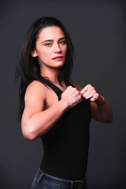 A woman poses confidently, adjusting her black tank top while standing against a neutral background. Her expression reflects determination and strength, highlighting her physique.