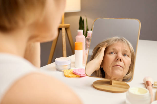 istock A senior woman carefully studies her reflection in a mirror while adjusting her hair. Various skincare products are neatly arranged on the vanity, creating a serene atmosphere 2201711544