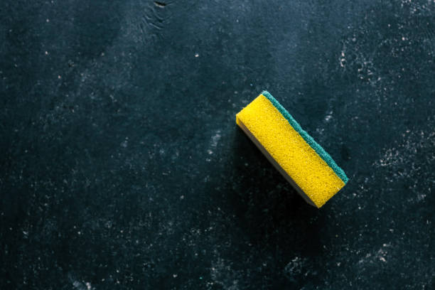 Single yellow dishwashing sponge with a green scrubbing layer placed on a dark textured background. Household cleaning tool for washing dishes, scrubbing surfaces, and general cleaning tasks.