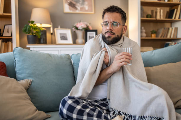 istock Sick man sitting on sofa, wrapped in blanket, holding glass of water 2201727095