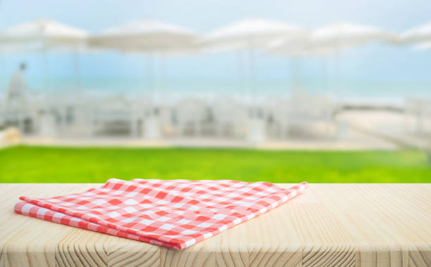 Red and white checkered cloth neatly placed on a wooden table with a blurred seaside resort in the background.For food and drink design ads with summer vacation themes.not ai generated