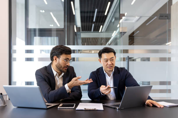 istock Two business colleagues discussing financial project while sitting at desk inside office, diverse people. Men serious and concentrated with laptops 2201736905