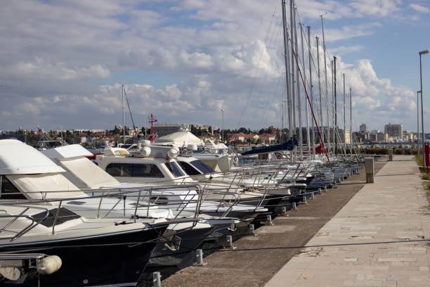A marina with yachts docked on a sunny day, featuring a cityscape in the background under a partly cloudy sky.