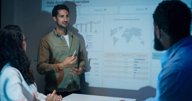 istock Group of businesspeople working late analyzing financial data displayed on projection screen while discussing strategy and planning for future business growth at office night. Finance meeting. 2201741570