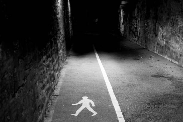 A pedestrian walkway, marked with a white line and a walking figure symbol, leads into a dark and narrow alleyway, suggesting a designated route for people to navigate through the urban space.