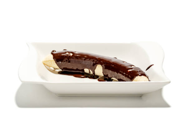 A banana covered in chocolate sauce. It is on a white plate. The plate is rectangular. The banana is on the left side of the plate