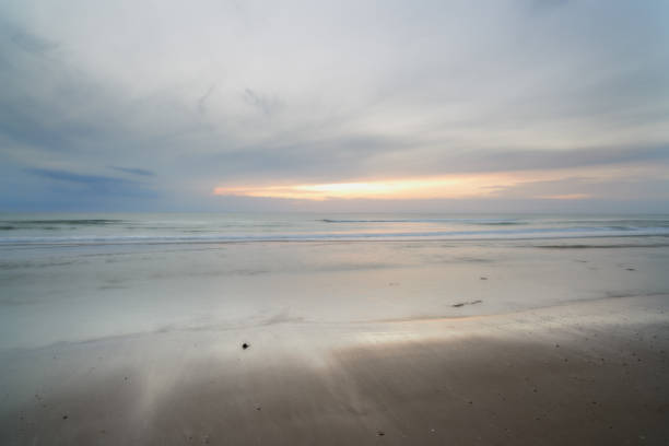 An inviting view of a peaceful coast under a cloudy sky with soft sunlight peeking through. The sea reflects the glowing hues while gentle waves create a soothing ambiance against the sandy shore.