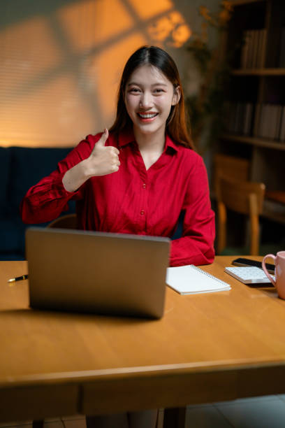 istock Female Asian professional giving thumbs up while working remotely at night, using laptop, notebook, smartphone, calculator on workspace desk 2201753984