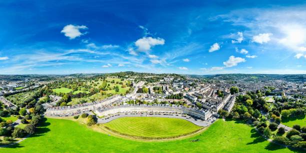 Aerial panoramic view of the Royal Crescent in Bath, England, showcasing Georgian architecture and lush greenery under a vibrant blue sky.