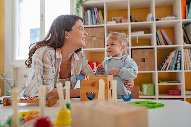 A young woman and a little girl play with educational toys at a preschool or daycare, engaging in learning activities with a shape sorter in a bright, cheerful classroom stock photo