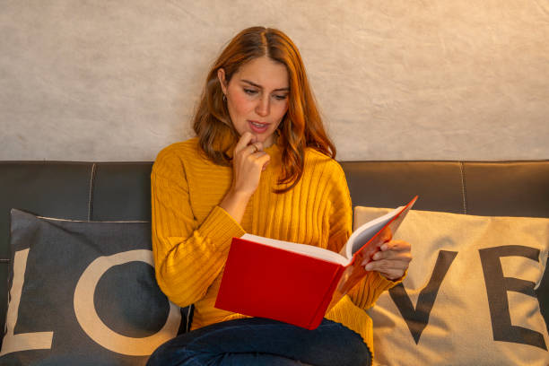 Young woman wearing a yellow sweater thoughtfully reading a red book while sitting on a comfortable sofa with love pillows