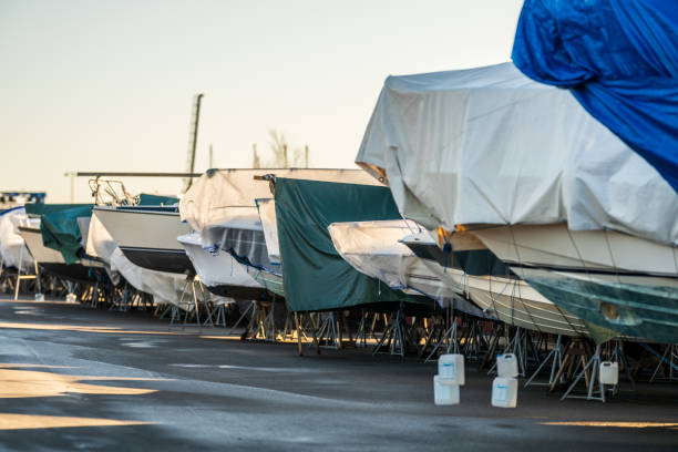 White and blue motor yacht with red canopy stored on supports in snowy winter boatyard. Pleasure boats on maintenance stands with snow covered ground clear blue sky boat storage facility