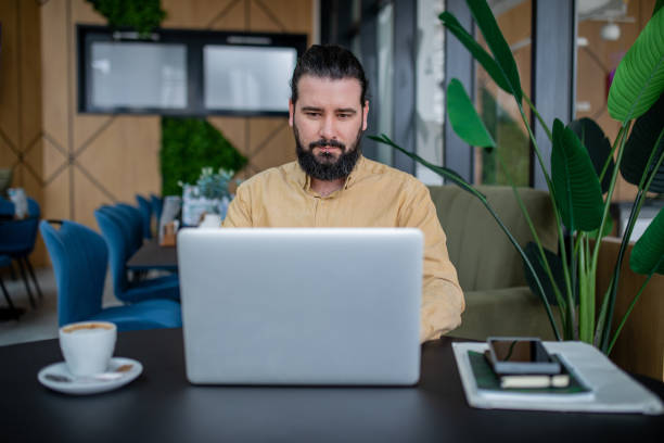 istock Front view of young, bearded man who is working on a laptop in coffee bar. 2201781193