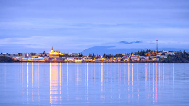 The town of Borgarnes reflected on still water at dusk in Iceland, with a church on a hill and mountains in the distance.