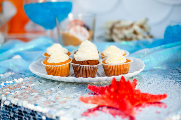 Vanilla cupcackes with white creamcheese on seashell shape plate. Silver fabric on the background.