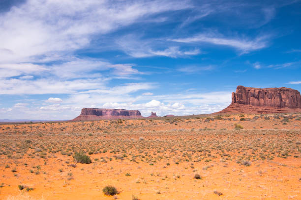 Navajo Tribal Park in Monumental Valley located in the Northern part of New Mexico. On a beautiful summer day, with towering red sandstone buttes rising dramatically from the vast desert floor. The deep blue sky, painted with wisps of white clouds, stretches endlessly over the iconic red rock formations.