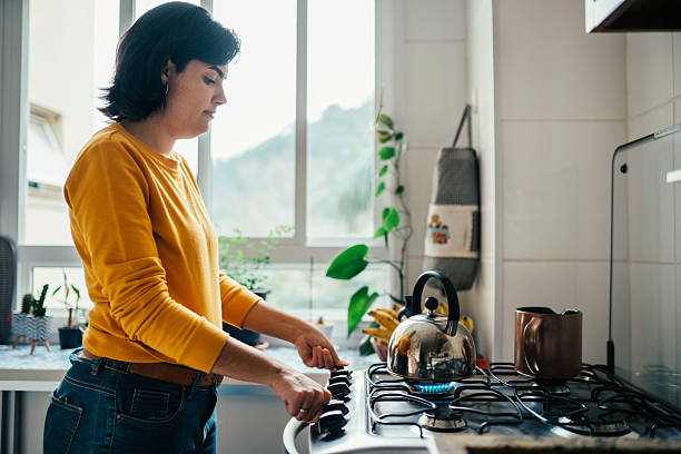Side view of young woman turning on the stove to boil water in the kettle to make tea in her kitchen