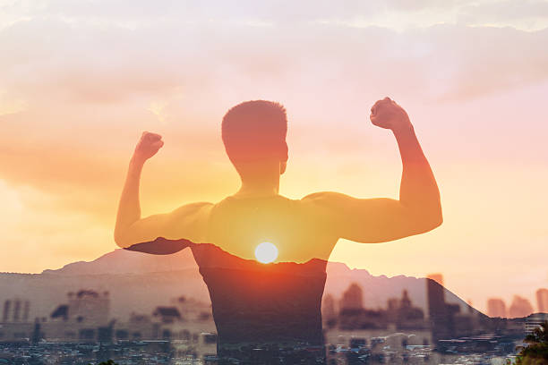 istock Man flexing muscles and looks out over the city skyline at sunset visualizing success. Strong person silhouette facing city, people reaching goals, having a vision, never give up concept. 2201809680