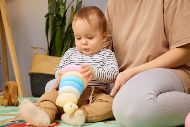 istock A young child sits on a play mat, focused on stacking colorful rings while a caregiver sits nearby. The cozy indoor setting features plants and wooden toys, creating a nurturing environment 2201812394