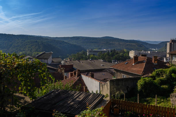 Serene village rooftops bask in sunlight, framed by lush green hills and distant buildings on a beautiful day.