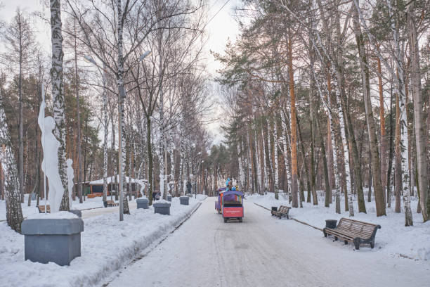 Yekaterinburg, Russia - February 14, 2025: Scenic winter park alley covered in snow, Soviet-era sculptures and tall birch trees. Colorful children's train moves along pathway. Serene, icy landscape.