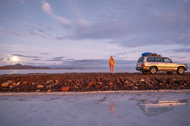Adventurous traveler stands by SUV on rocky terrain. Scenic sunrise landscape with rugged 4x4. Solitary journey in remote desert region evokes wanderlust.