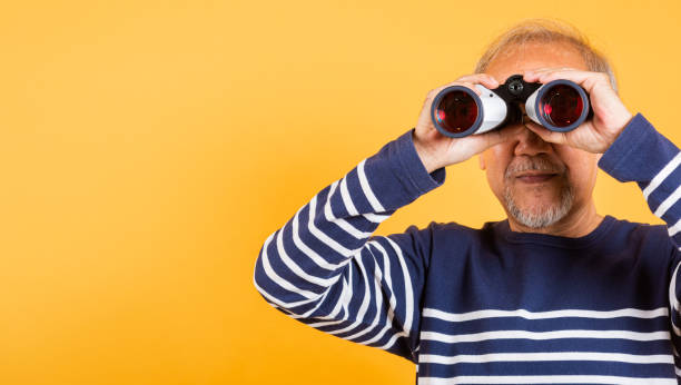 istock Portrait Asian smiling old man looking camera through binoculars studio shot isolated yellow background, senior man pensioner feeling surprised 2201828129