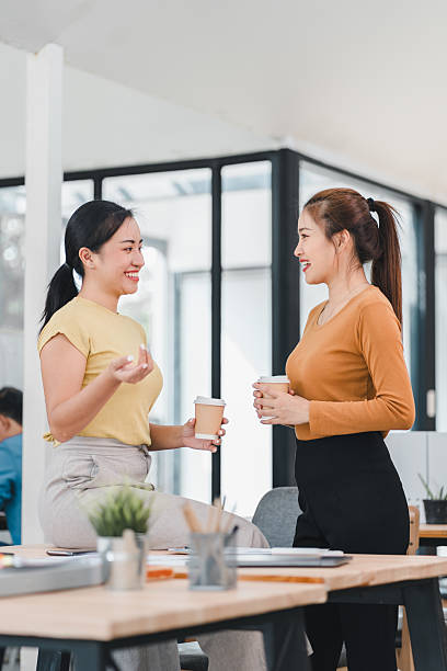 istock Two women talking and smiling while holding coffee cups in office 2201828241
