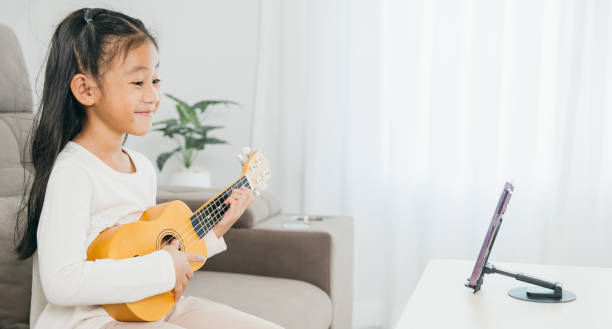 istock A young girl sits on a couch playing a ukulele, with a smartphone on a stand streaming her online music lesson. The scene highlights creative learning at home 2201828552