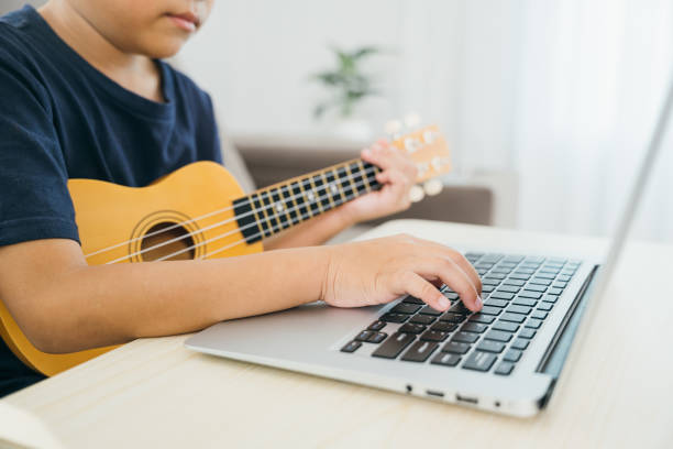 istock Close-up of a young boy hands strumming a ukulele while watching an online tutorial on a laptop. Sitting on the sofa in the living room, he blends traditional music practice with modern learning 2201828559