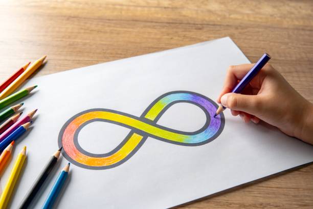 A hand coloring a rainbow infinity symbol on a white sheet of paper with colored pencils scattered around.