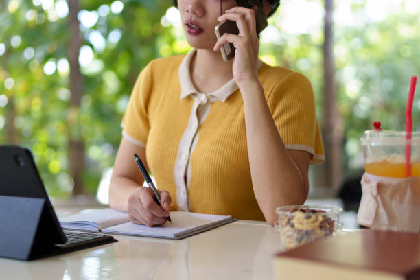 istock Young Asian woman talking on the phone while taking notes and studying in a university cafe with books and a laptop on the table 2201844759