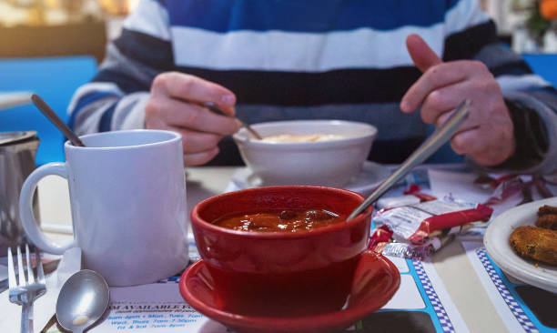 54-year-old man enjoys a bowl of pureed soup in a classic American small-town diner. In the foreground, a red bowl filled with chili soup sits on the table, adding warmth and depth to the cozy dining scene.
