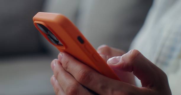 Close-up of a mobile phone with three cameras, in an orange case in men's hands. Man typing on smartphone, no face