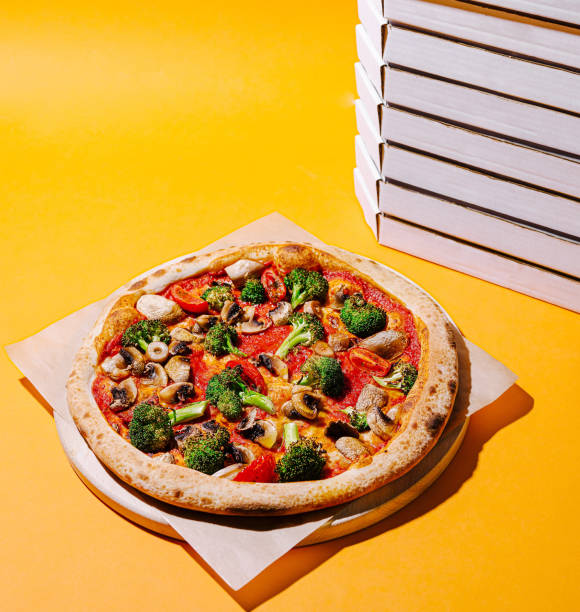 istock Colorful vegetable pizza placed on a wooden board beside stacked pizza boxes against a bright yellow background 2201852232