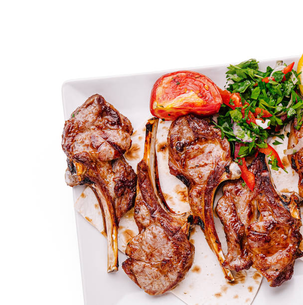 Juicy grilled lamb chops are served on a white plate alongside a vibrant salad of fresh herbs and a roasted tomato, showcasing a colorful and appetizing dish.