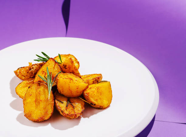 Crispy roasted potatoes served on a white plate, garnished with fresh rosemary, against a vibrant purple background. Perfectly golden and inviting, ideal for any meal.