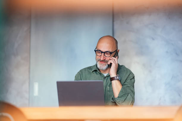 Mature bald man smiling while speaking on a smartphone, working on a laptop indoors, representing productivity and modern communication in a coworking space.