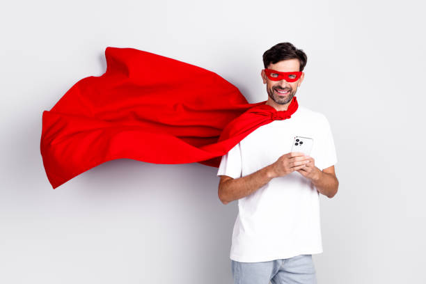 istock Happy young man dressed as superhero with red cape and mask holding a smartphone, standing on white background 2201864311