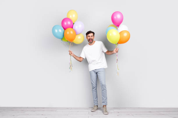 istock Cheerful young man holding colorful balloons in both hands on a light gray background, showcasing joy and celebration in casual outfit. 2201864326