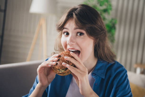 istock Young woman enjoying a delicious burger at home with a cheerful expression while relaxing in a modern cozy living room 2201865189